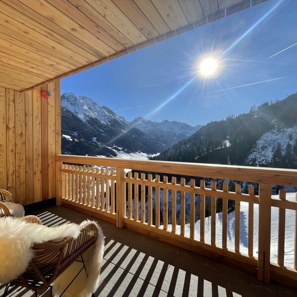 Sonniger Balkon der Lodge mit Holzelementen, zwei Stühlen mit Fellen und Blick auf die schneebedeckten Berge.