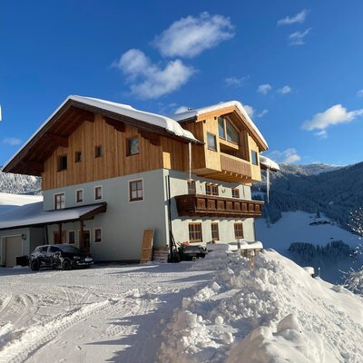 Die winterliche Außenansicht des Bio-Bauernhofs, umgeben von Schnee und Bergen unter blauem Himmel.