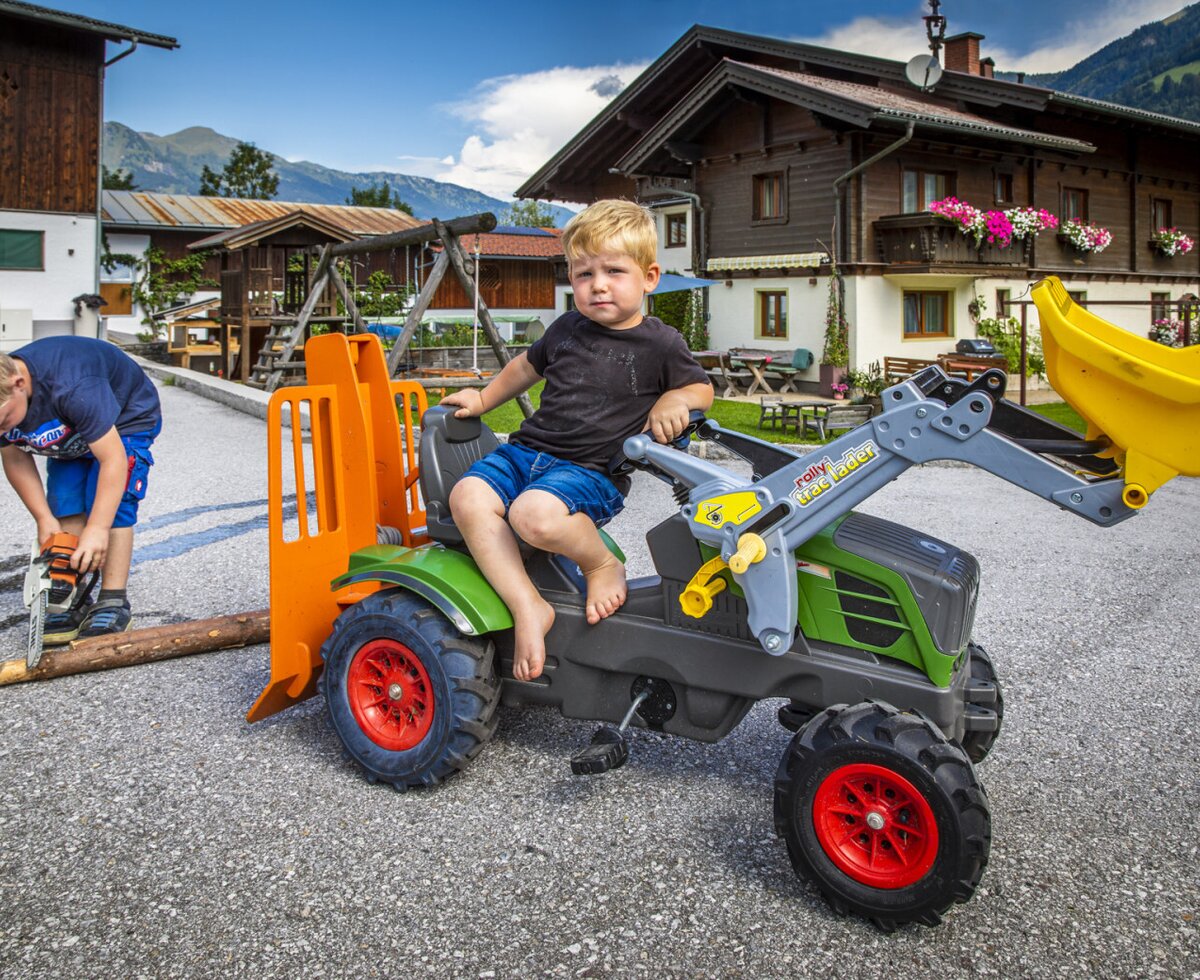 Kinder spielen auf dem Bauernhof mit einem Trettraktor und einer Spielzeugkettensäge, wobei ein Spielplatz im Hintergrund sichtbar ist.