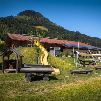 Der einladende Spielplatz des Bauernhofs mit einer gelben Rutsche, Holzbänken und einer Spielhütte vor malerischer Bergkulisse.