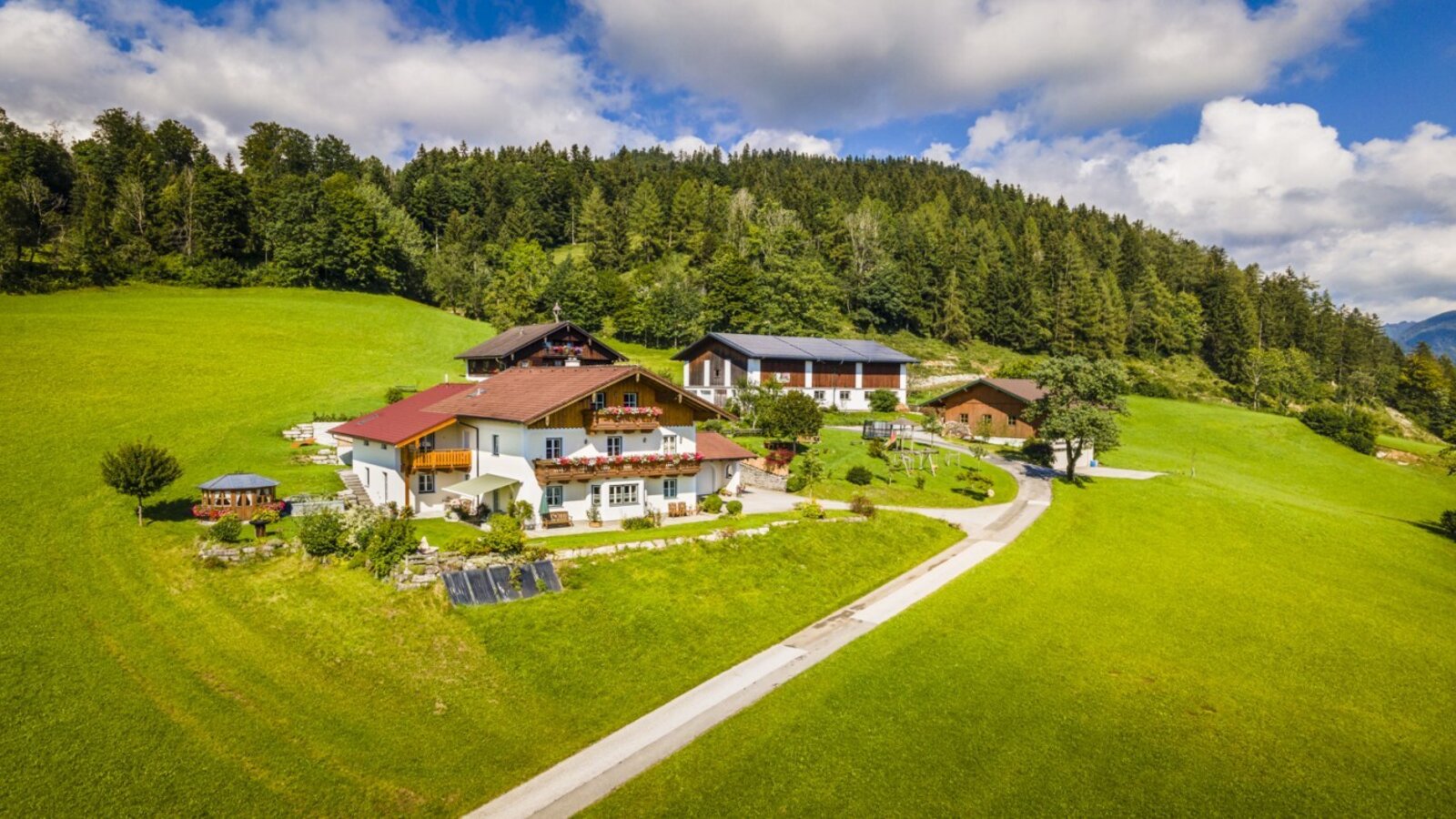 Der Bauernhof mit seinen Gebäuden liegt inmitten grüner Wiesen und Wälder und bietet einen Blick auf die umliegenden Berge.