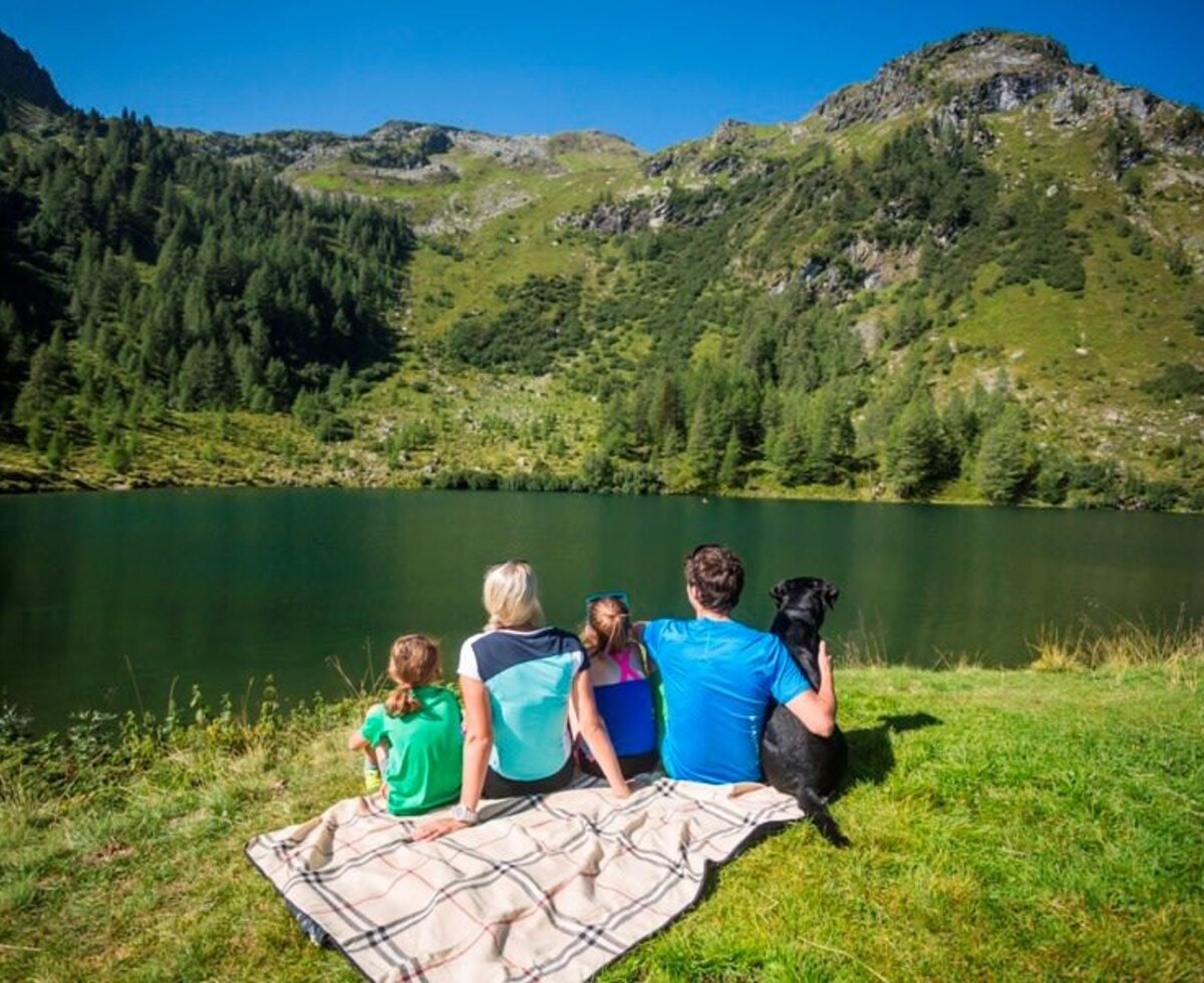 Familie und Hund sitzen am Ufer eines Bergsees mit Blick auf die bewaldete Berglandschaft.