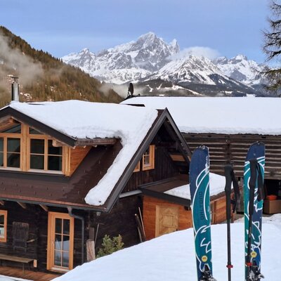 Unsere Alm im Winter, mit schneebedecktem Dach, Bergblick und Skiern im Vordergrund.