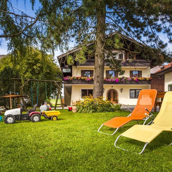 Der Garten des Bauernhofs mit einer Rasenfläche, zwei Sonnenliegen, einer Schaukel und einem Spieltraktor für Kinder.