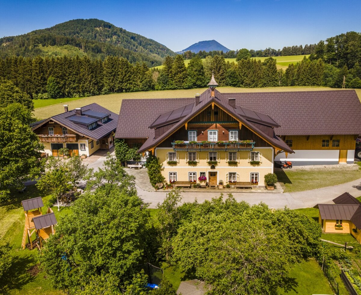 Der Bauernhof mit seinen traditionellen Gebäuden, einem Gartenbereich und der umliegenden Landschaft mit Wiesen und bewaldeten Bergen.