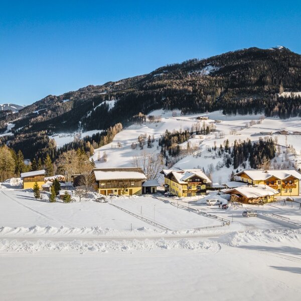 Der Bauernhof in einer sonnigen Winterlandschaft, umgeben von schneebedeckten Bergen und Wäldern, mit Langlaufloipen im Vordergrund.