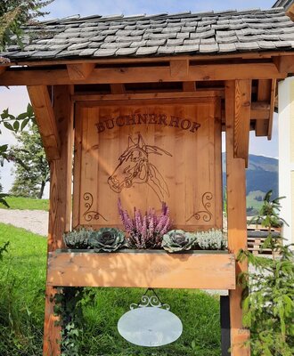 Hölzernes Schild des Bauernhofs "Buchnerhof" mit geschnitztem Pferdemotiv und dekorativen Pflanzen.
