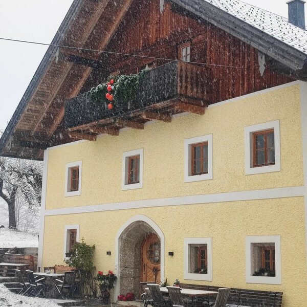 Außenansicht des Bauernhofs im Schnee, mit gelber Fassade, Holzbalkon und Sitzgelegenheiten im Freien.