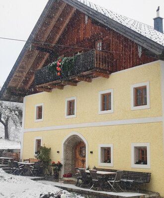 Außenansicht des Bauernhofs im Schnee, mit gelber Fassade, Holzbalkon und Sitzgelegenheiten im Freien.