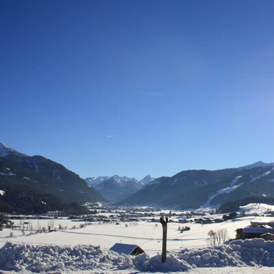 Blick auf die verschneite Berglandschaft mit einem Tal, einem Dorf und Skipisten bei Sonnenschein.