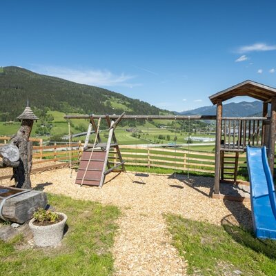 Kinderspielplatz des Bed and Breakfast mit Rutsche, Schaukel und Wasserspiel, umgeben von einem Holzzaun und mit Bergblick.