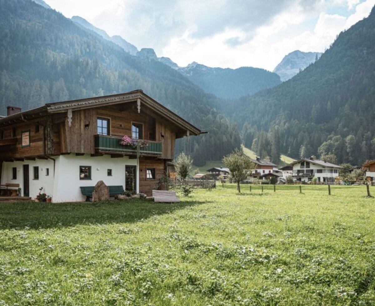 Außenansicht des Bed and Breakfast mit traditioneller Holzfassade, Balkon und umliegender Wiese vor einer Bergkulisse.