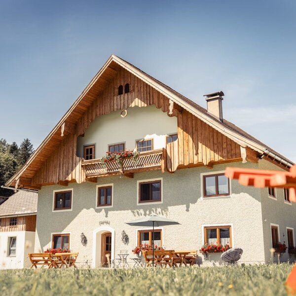 Der Bauernhof von außen, ausgestattet mit einem Balkon, Blumenkästen und einem Sitzbereich mit Tischen und Bänken.
