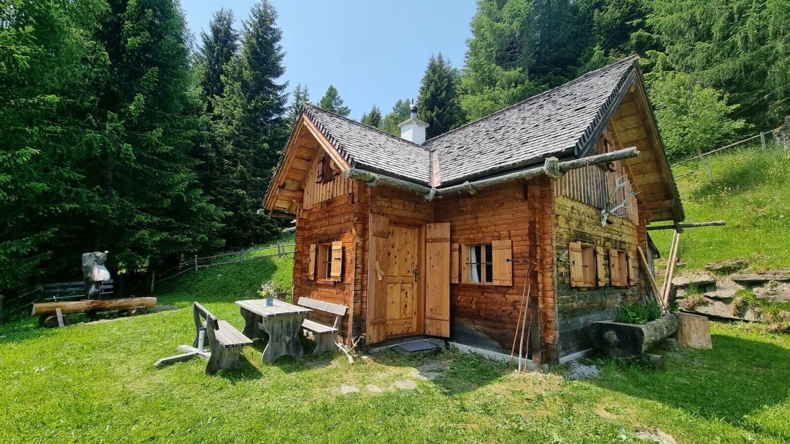 Die Almhütte von außen, umgeben von Wald und Wiesen, mit einem Essbereich im Freien.