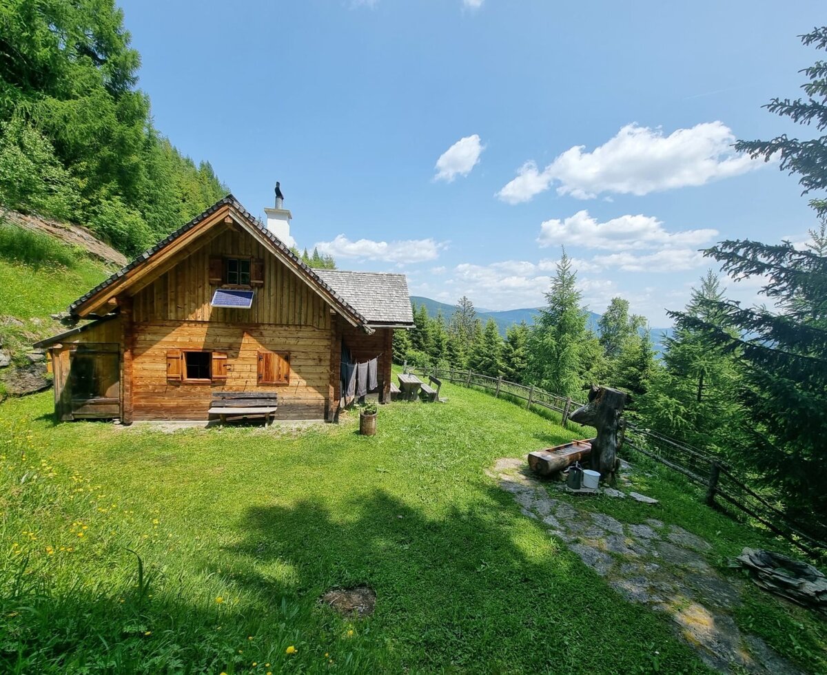 Die Almhütte mit Solarpaneel und Außensitzgelegenheiten, umgeben von Wiesen und Bergen.