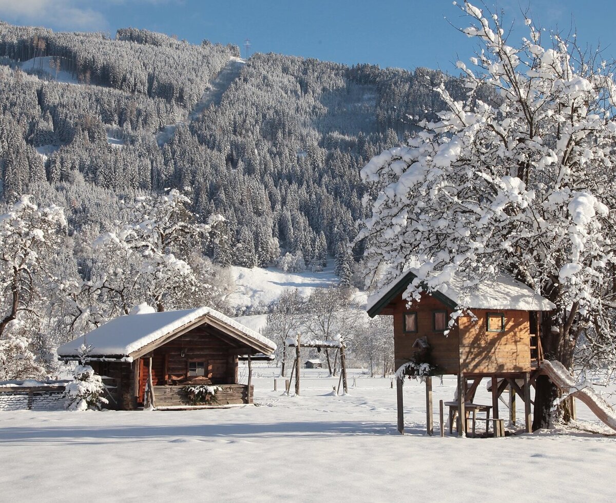 Schneebedeckter Gartenbereich des Bauernhofs mit einem Holzspielhaus, Rutsche und Schaukeln vor einer bewaldeten Bergkulisse.