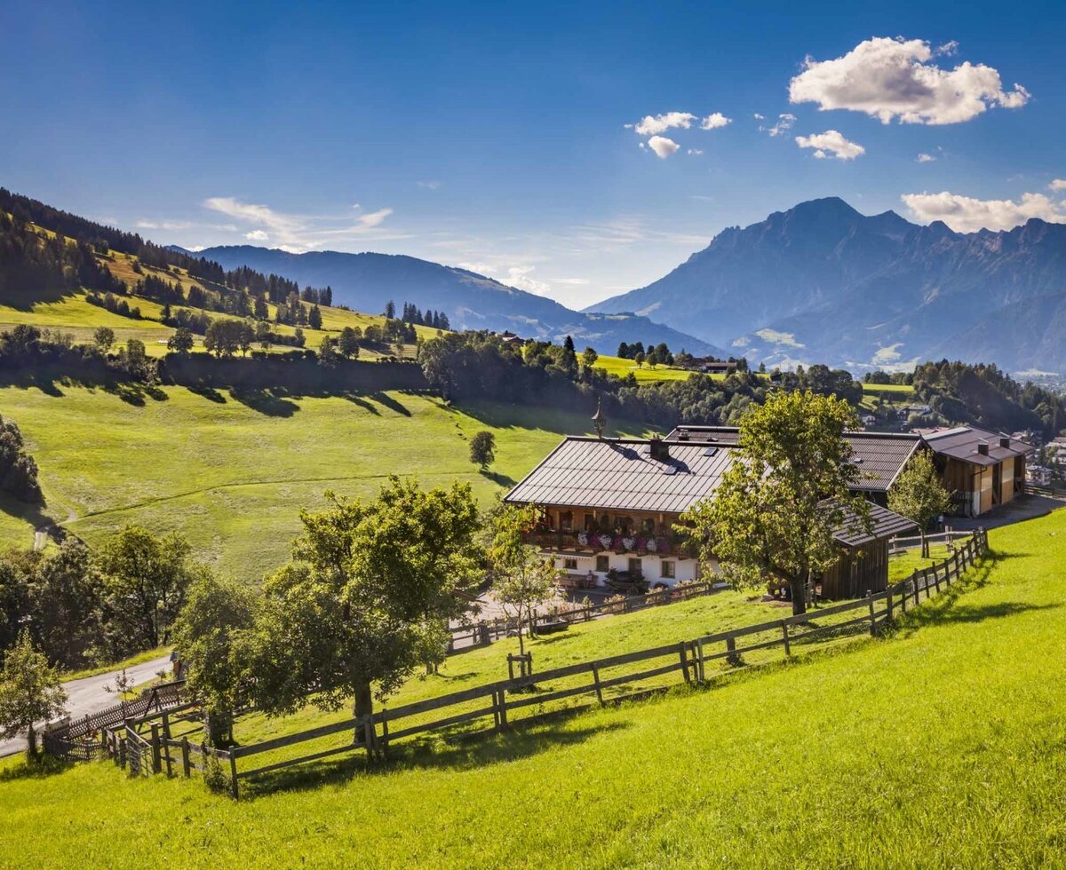 Der Bauernhof in einer weitläufigen Berglandschaft, umgeben von grünen Wiesen und Bergen.