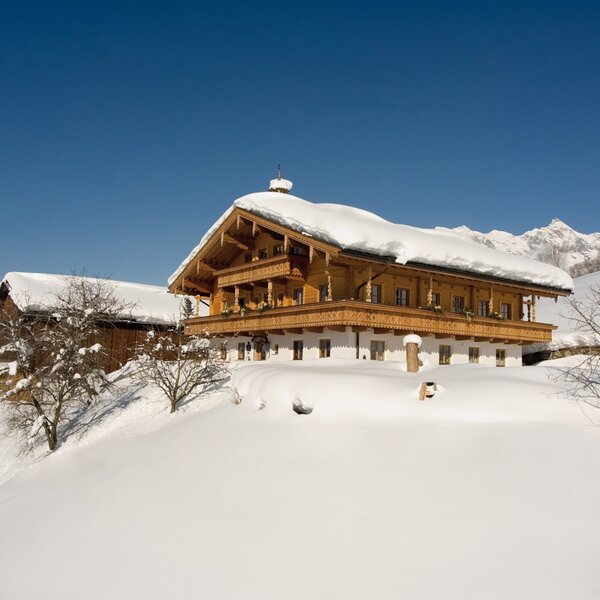 Der Bauernhof mit Holzbalkonen und schneebedecktem Dach, eingebettet in eine winterliche Berglandschaft.