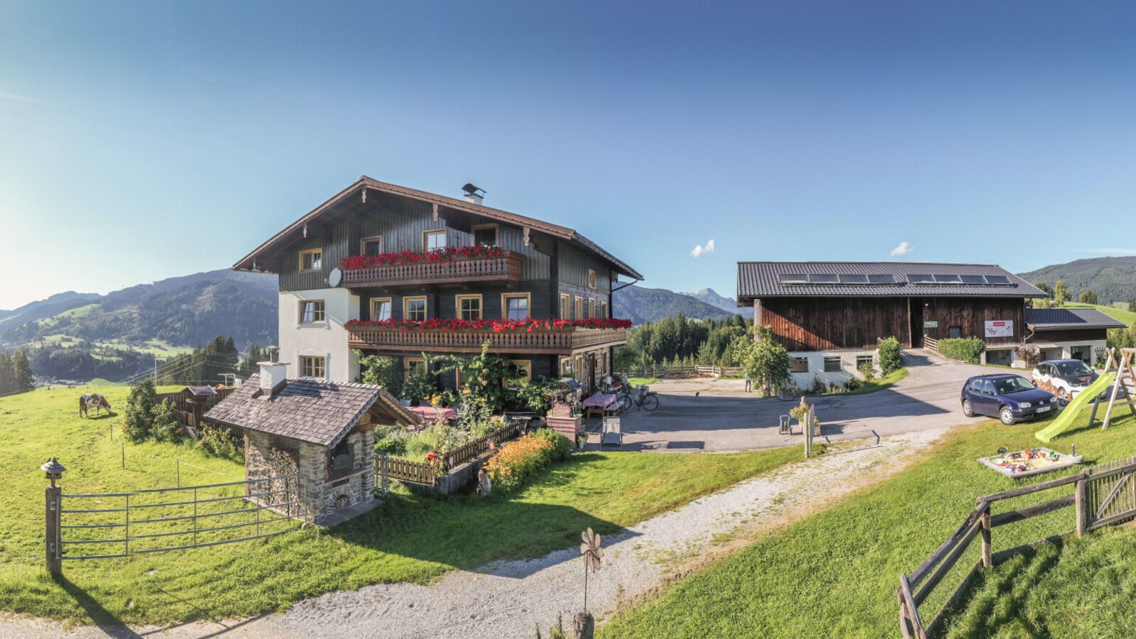 Der Bauernhof mit dem Haupthaus, Balkonen mit Blumen, einem Wirtschaftsgebäude, Kinderspielplatz und umliegenden Wiesen mit Bergblick.