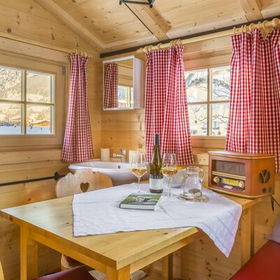 Essbereich in der Auszeithütte mit Holzwänden, einem Tisch für zwei Personen, Spülbecken, Fenstern mit karierten Vorhängen und einem Radio.