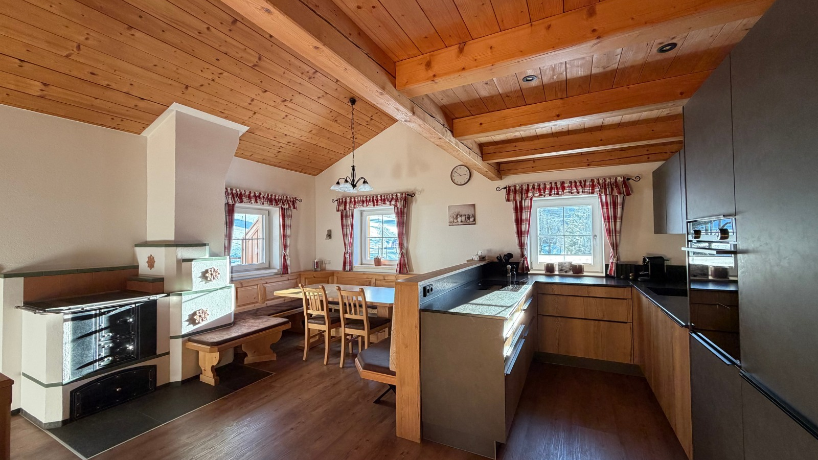 Moderne Küche und Essbereich der Alm mit Holztisch, Bänken, traditionellem Kachelofen und Holzbalkendecke.