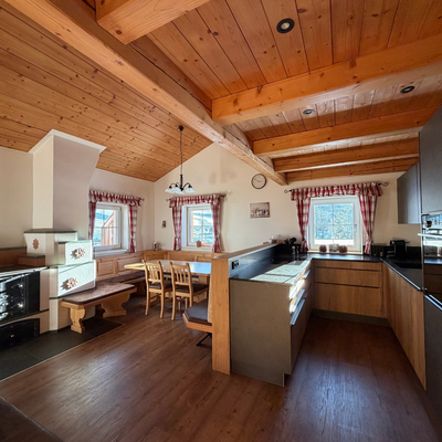 Moderne Küche und Essbereich der Alm mit Holztisch, Bänken, traditionellem Kachelofen und Holzbalkendecke.