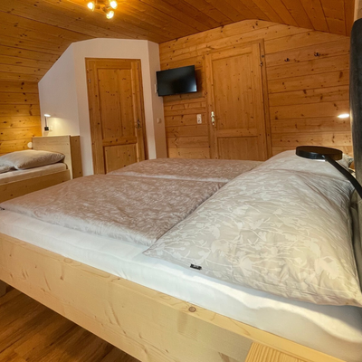 Schlafzimmer in der Alm mit einem Doppelbett, einem Einzelbett, Holzwänden, einem Fernseher und Leselampen.