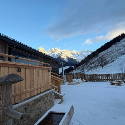 Die Alm mit ihrer Holzfassade und dem verschneiten Außenbereich mit Holzbänken und Bergblick.