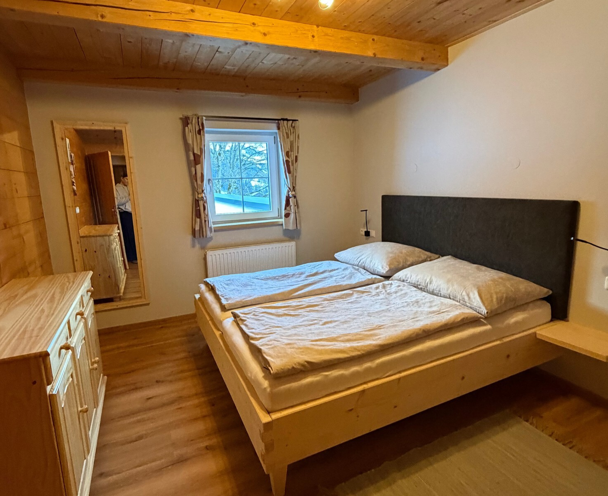 Schlafzimmer der Alm mit Doppelbett, Holzmöbeln, Fenster und Holzbalkendecke.