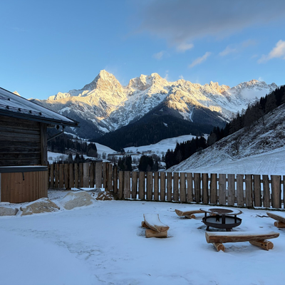Verschneiter Außenbereich der Alm mit Hot Tub, Feuerstelle, Holzbänken und Panoramablick auf die Berge.