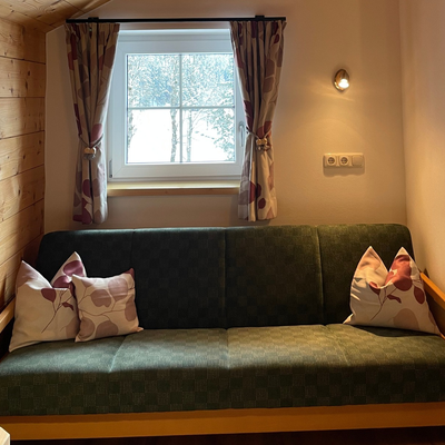 Schlafsofa in der Alm mit Holzwand, Fenster und Vorhängen, das zusätzlichen Schlafplatz bietet.