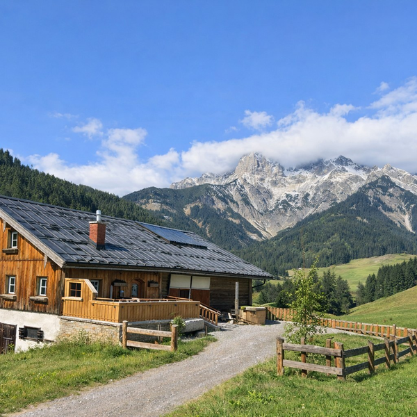 Die Alm mit Holzfassade, dunklem Dach und Solarpaneelen, gelegen an einem Schotterweg mit Holzzaun, vor einer Kulisse aus grünen Wiesen und schneebedeckten Bergen.