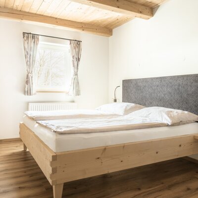 Schlafzimmer der Alm mit Doppelbett, Holzrahmen, grauer Kopflehne, integrierten Nachttischen, Leselampen, Fenster, Holzdecke und einem Spiegel.