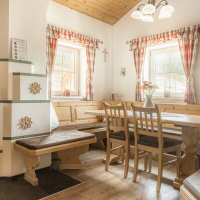 Essbereich der Alm mit Kachelofen, Holztisch, Stühlen und einer Eckbank.