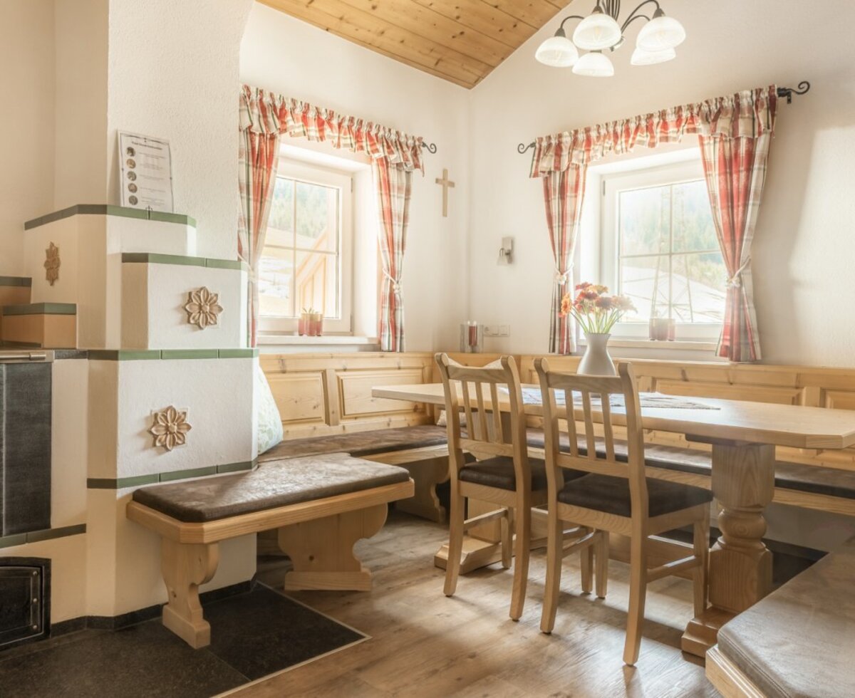 Essbereich der Alm mit Kachelofen, Holztisch, Stühlen und einer Eckbank.