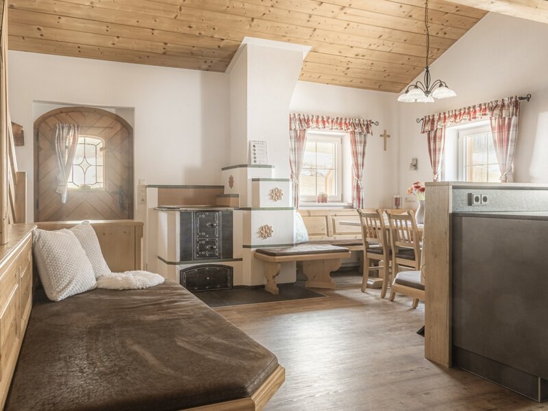 Wohnbereich der Alm mit Tagesbett, traditionellem Kachelofen und Essbereich.