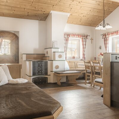 Wohnbereich der Alm mit Tagesbett, traditionellem Kachelofen und Essbereich.