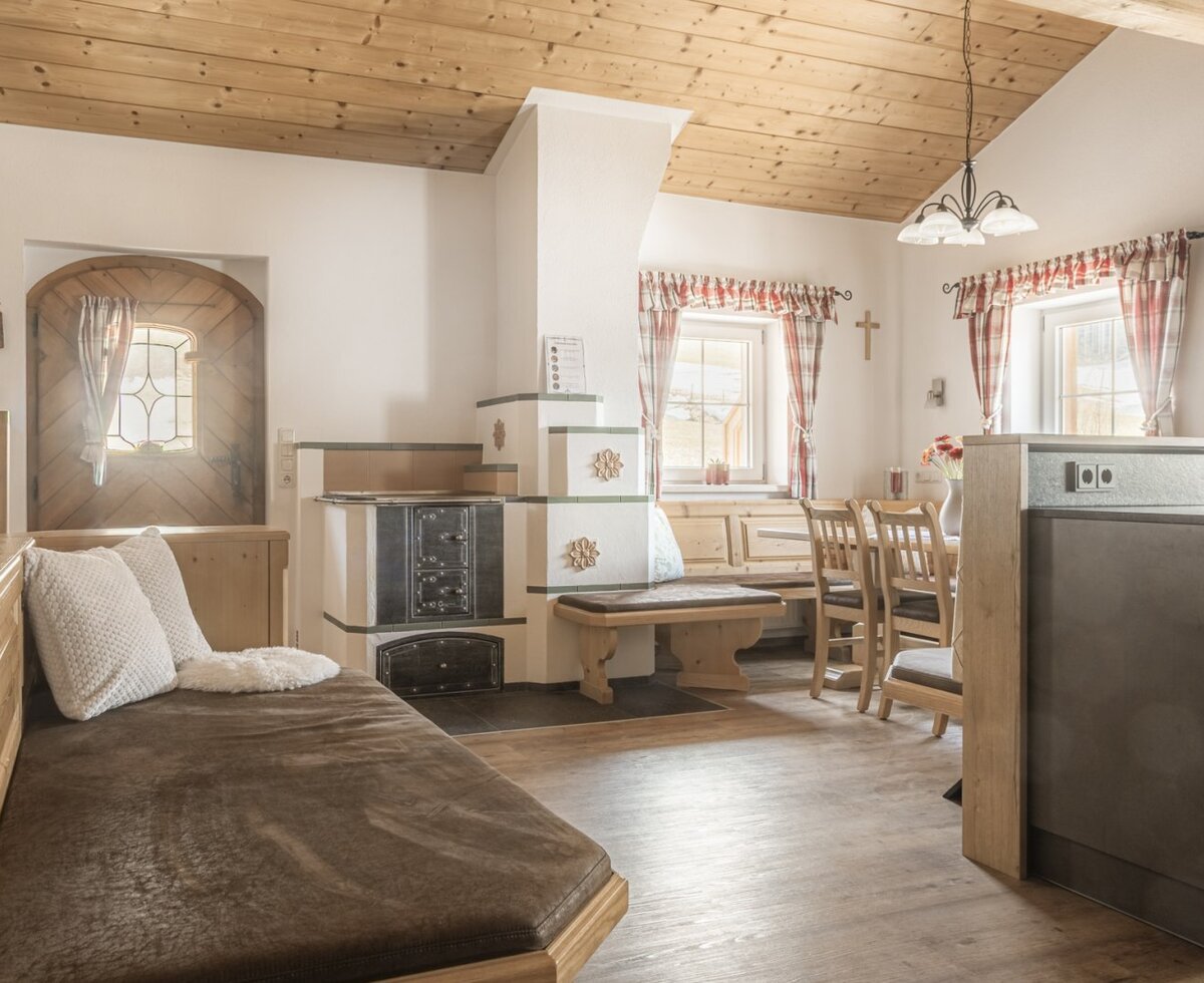Wohnbereich der Alm mit Tagesbett, traditionellem Kachelofen und Essbereich.