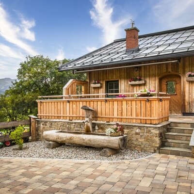 Die einladende Almhütte mit traditioneller Holzfassade, einer gemütlichen Holzterrasse und einem urigen Brunnen im gepflasterten Eingangsbereich.