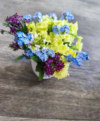 Ein kleiner Blumenstrauß mit gelben, blauen und violetten Blüten in einer weißen Vase auf einem Holztisch im Bauernhof.