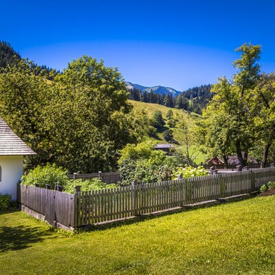 Eine kleine weiße Kapelle und ein umzäunter Garten mit Rasenfläche auf dem Bauernhof, umgeben von Hügeln und Bergen.