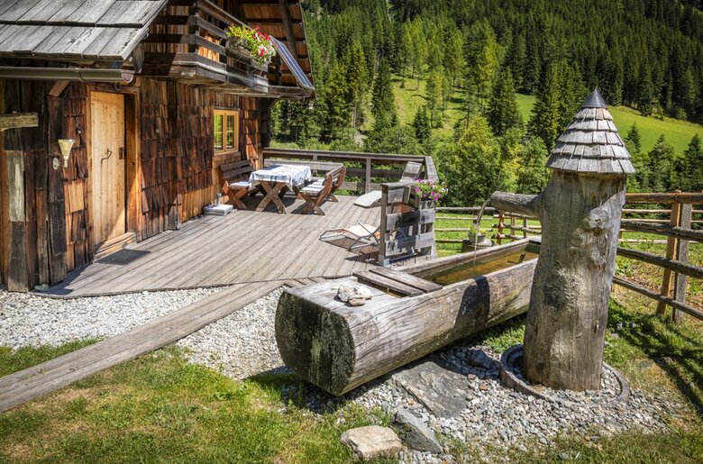 Die Holzterrasse des Hofs mit einem Esstisch und Bänken sowie einem Quellwasserbrunnen mit Holztrog.