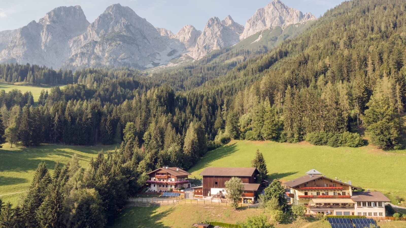 Der Bauernhof mit seinen Gebäuden liegt eingebettet in grüne Wiesen, umgeben von Wäldern und Bergen.