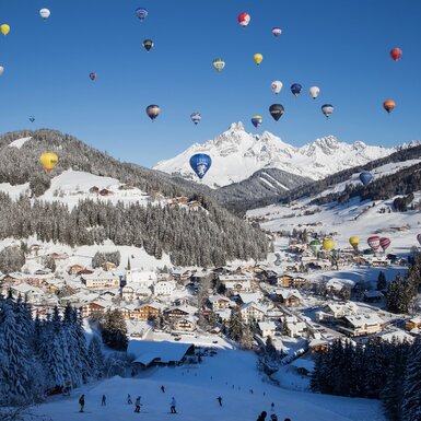 Blick auf die winterliche Landschaft von Filzmoos mit einem Skihang, dem Dorf und vielen bunten Heißluftballons am Himmel, die Gäste des Bauernhofs erleben können.
