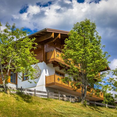 Die Außenansicht des Bauernhofs zeigt die Fassade mit Holzbalkonen und Bemalung auf einem Hanggrundstück.