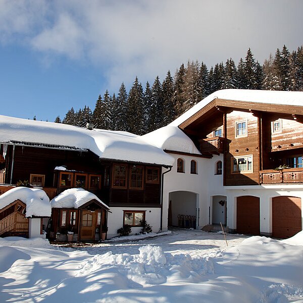 Außenansicht des schneebedeckten Bauernhofs mit traditioneller Holzfassade, Balkonen und Wald im Hintergrund.