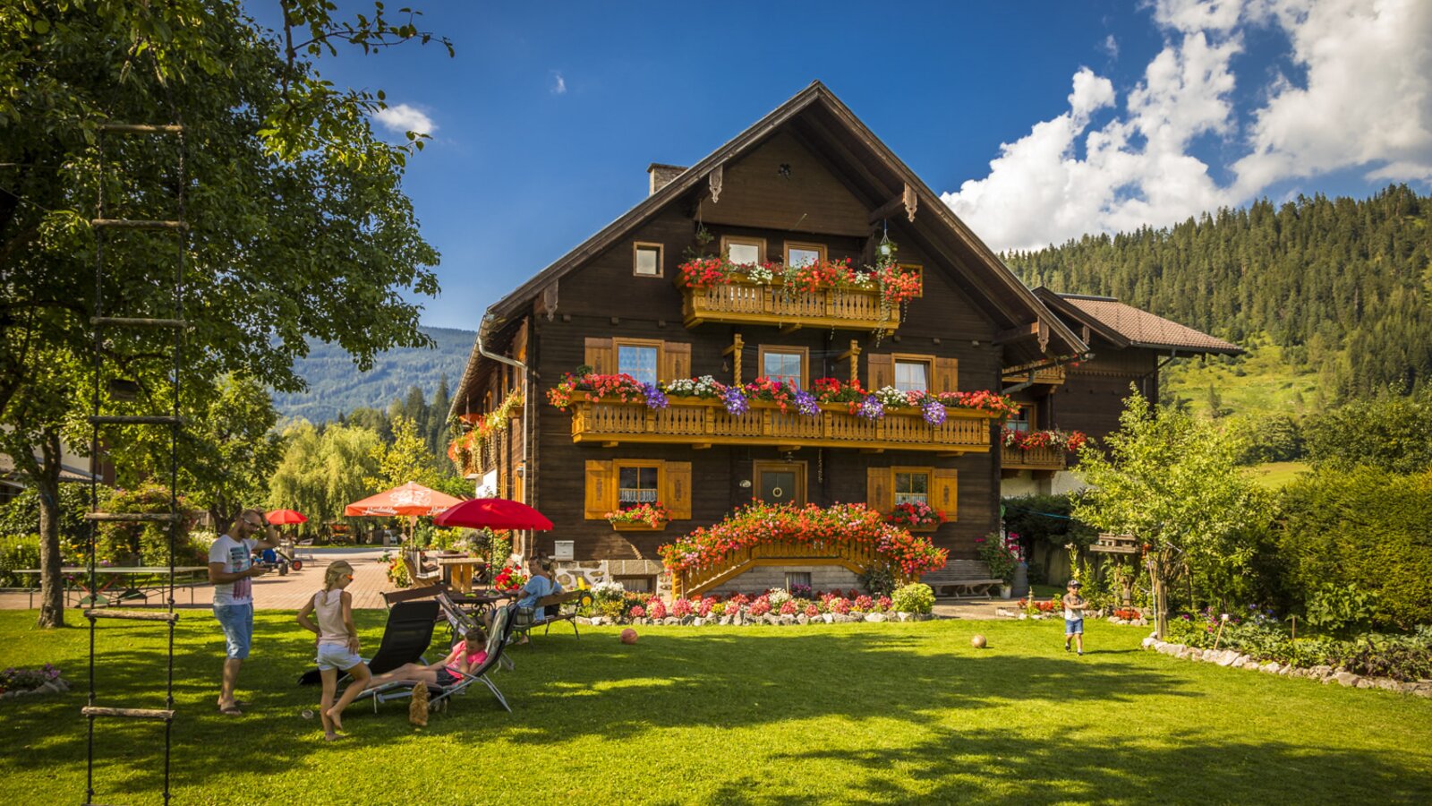 Der Bauernhof mit großem Garten inklusive Liegewiese und Spielbereich für Gäste, umgeben von blumengeschmückten Balkonen und der Berglandschaft.