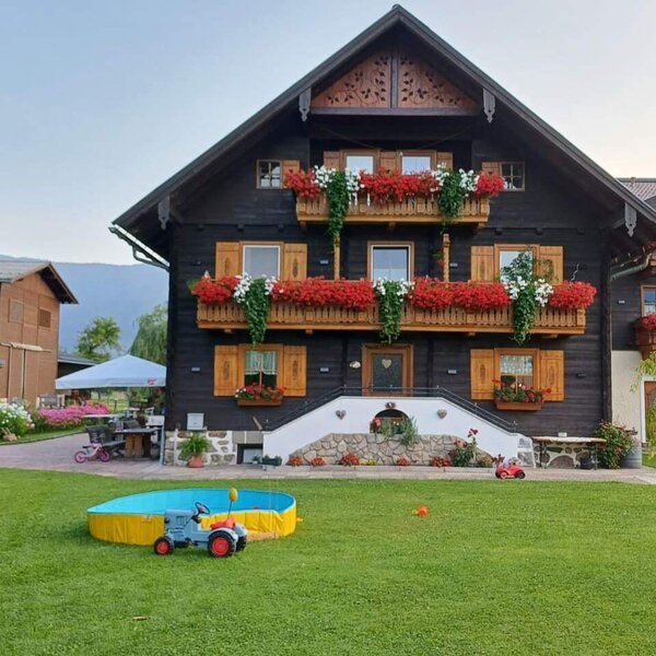 Außenansicht des Bauernhofs mit blumengeschmückten Balkonen und einem Garten mit Rasenfläche, Kinderplanschbecken und Spielzeugtraktor.