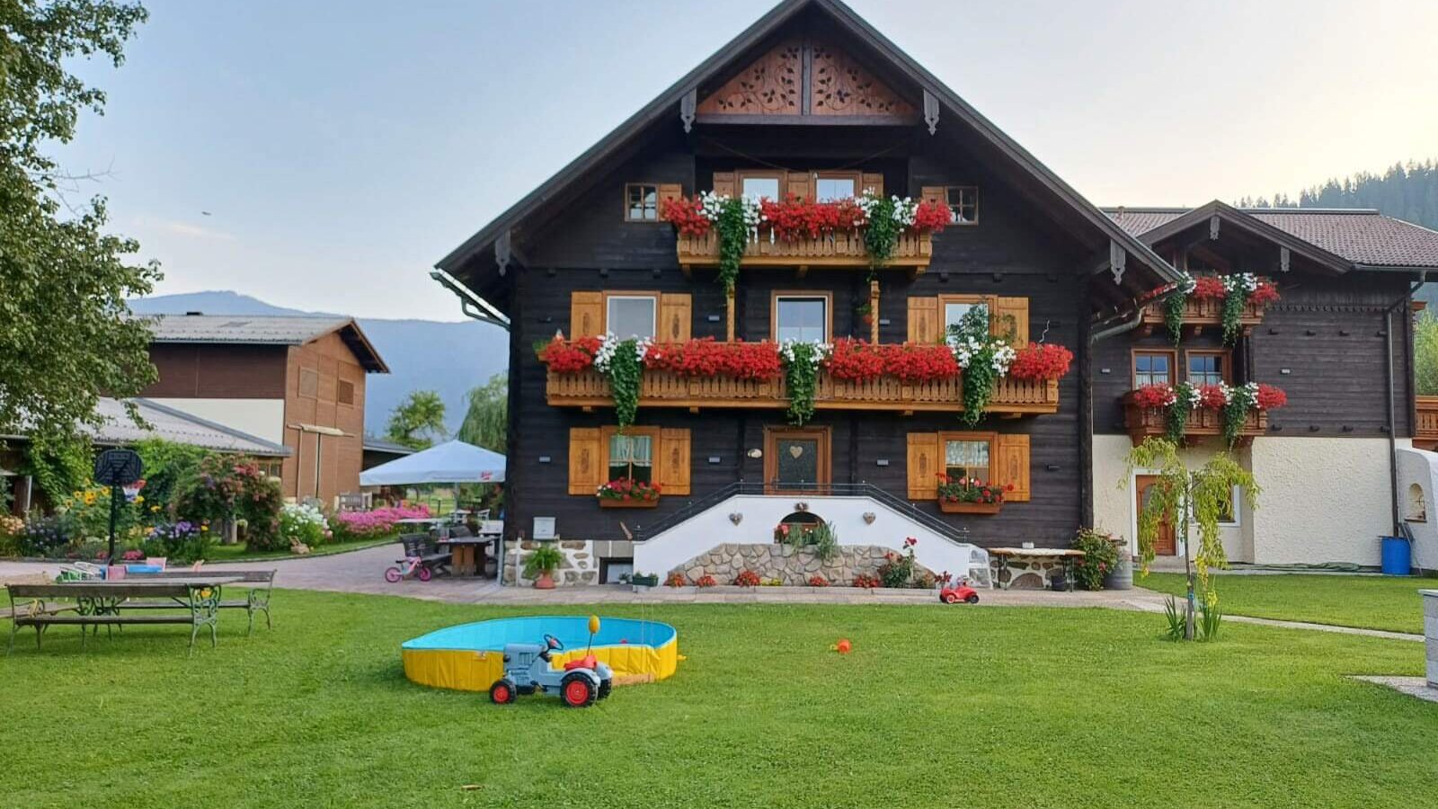 Außenansicht des Bauernhofs mit blumengeschmückten Balkonen und einem Garten mit Rasenfläche, Kinderplanschbecken und Spielzeugtraktor.