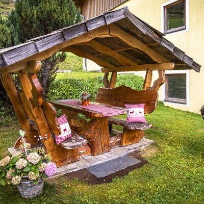 Ein einladender, überdachter Holzsitzplatz im Garten des Bauernhofs mit rustikalen Bänken, gemütlichen Kissen und einem bereitstehenden Kugelgrill.
